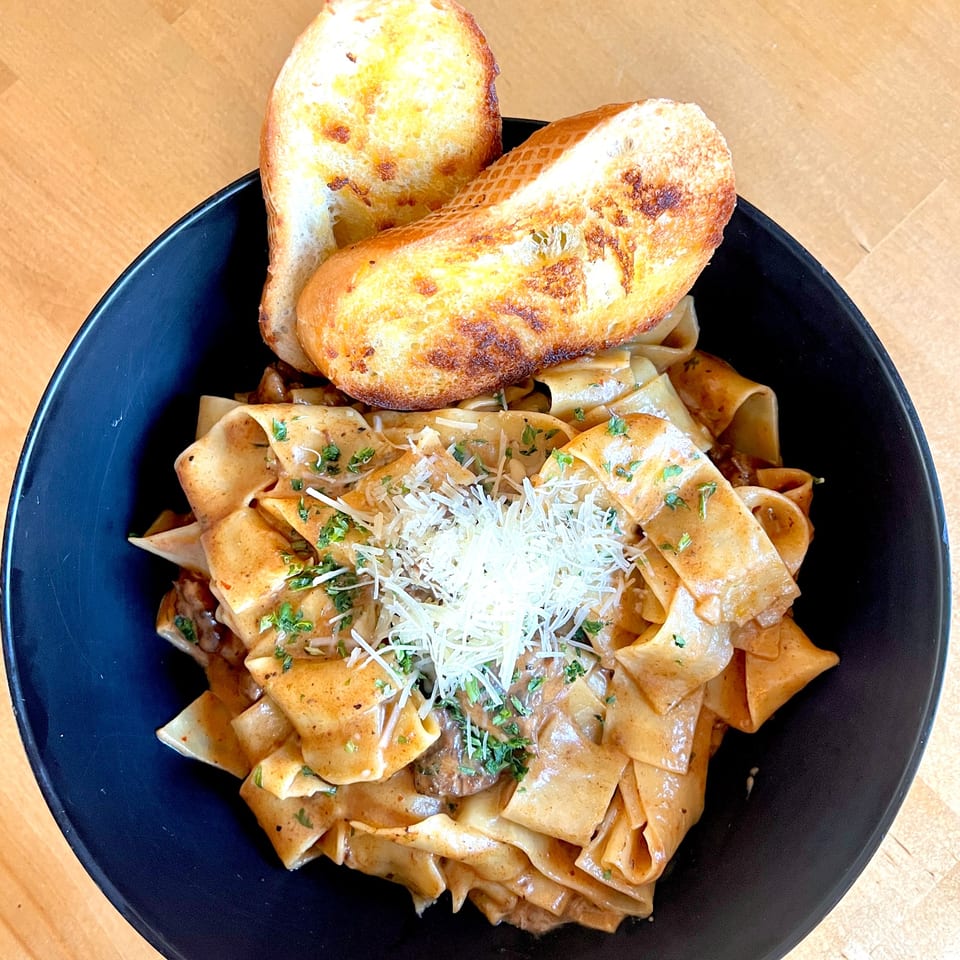 Pappardelle pasta with creamy sauce, grated cheese, parsley, and two slices of garlic bread in a black bowl.