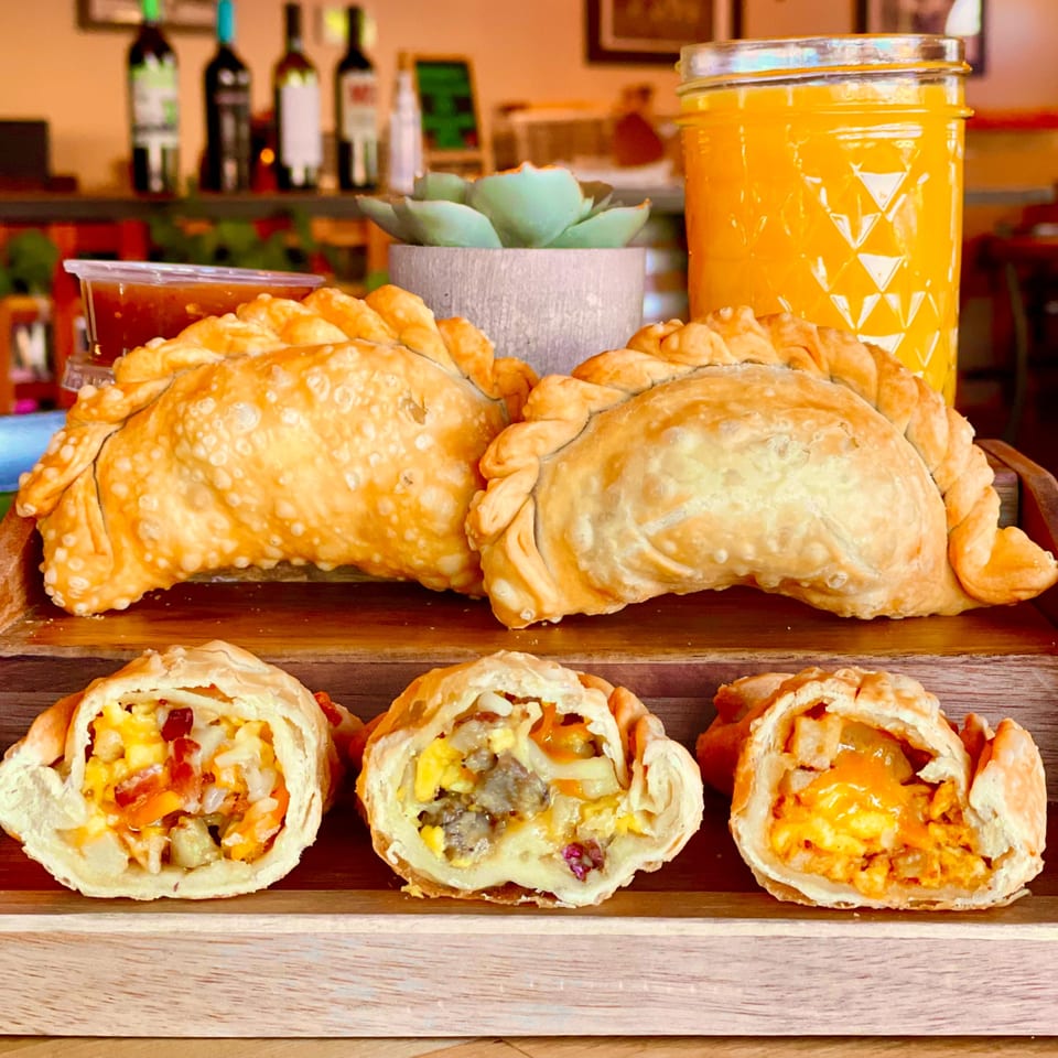 Lord Empanada's golden-brown empanadas, some cut open to show savory fillings, with a drink and sauce.