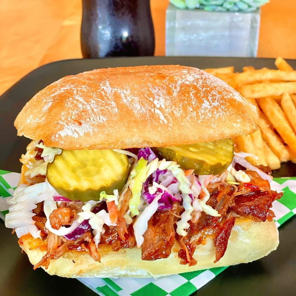 Pulled pork sandwich with coleslaw and pickles on a bun, served with a side of french fries.