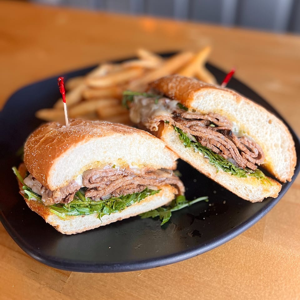 Two halves of a steak sandwich with arugula and onions, served with fries on a black plate.