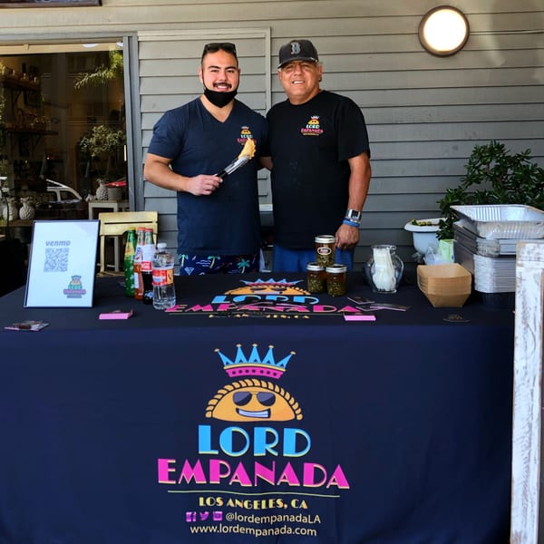 Two men, one holding an empanada with tongs, stand behind a Lord Empanada vendor table.