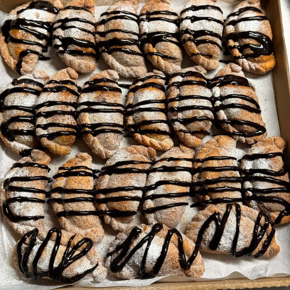 A box of sweet Argentinian empanadas, dusted with powdered sugar and drizzled with chocolate sauce.